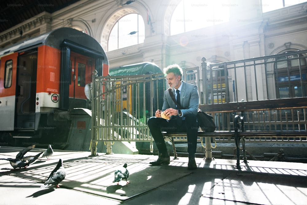 Belo empresário maduro em uma cidade. Um homem esperando o trem na estação ferroviária. Um homem sentado em um banco, alimentando pombos.