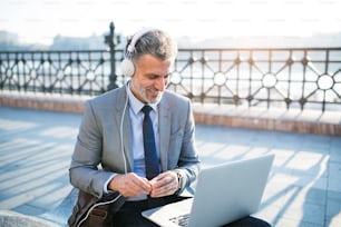 Handsome mature businessman with laptop in a city. Man sitting on a bridge, using laptop and headphones.