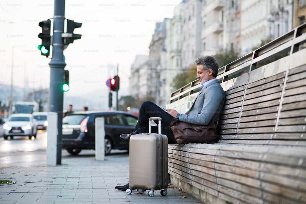 Bel homme d’affaires mature avec smartphone dans une ville, assis sur un banc, textant.
