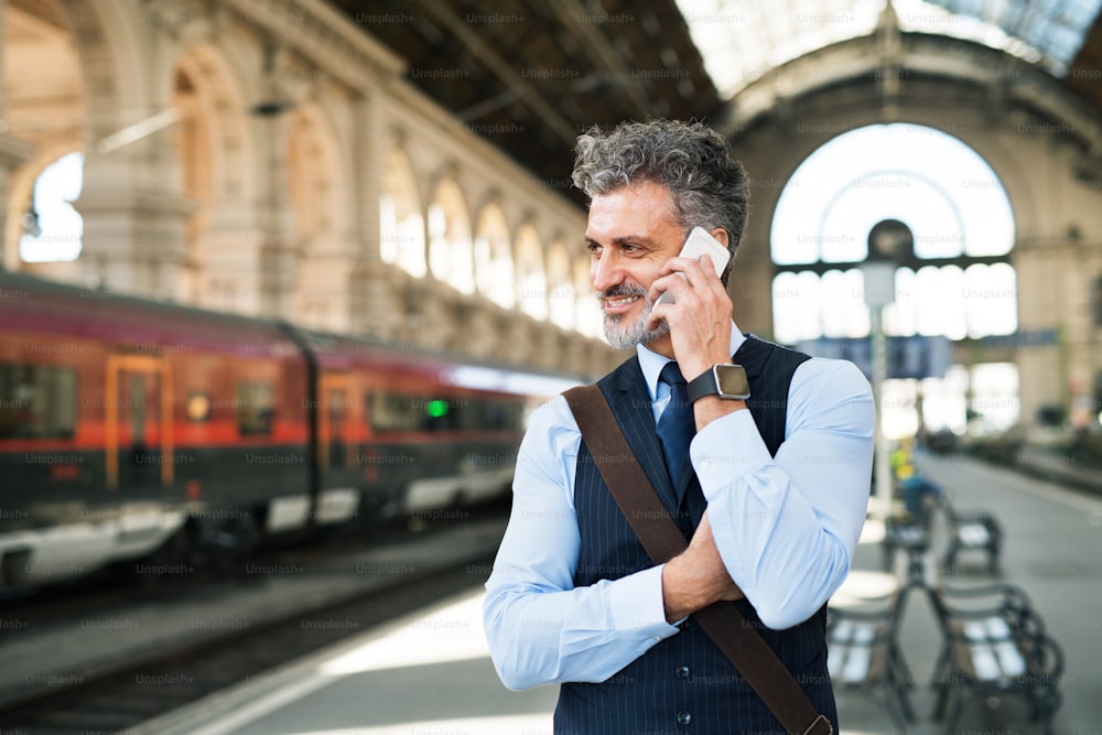 Bel homme d’affaires mature avec smartphone dans une ville. Homme attendant le train à la gare, passant un appel téléphonique.