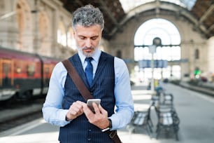 Gutaussehender reifer Geschäftsmann mit Smartphone in einer Stadt. Mann, der am Bahnhof auf den Zug wartet, SMS.