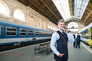 Gutaussehender reifer Geschäftsmann mit Smartphone in einer Stadt. Ein Mann, der am Bahnhof auf den Zug wartet und einen Anruf tätigt.