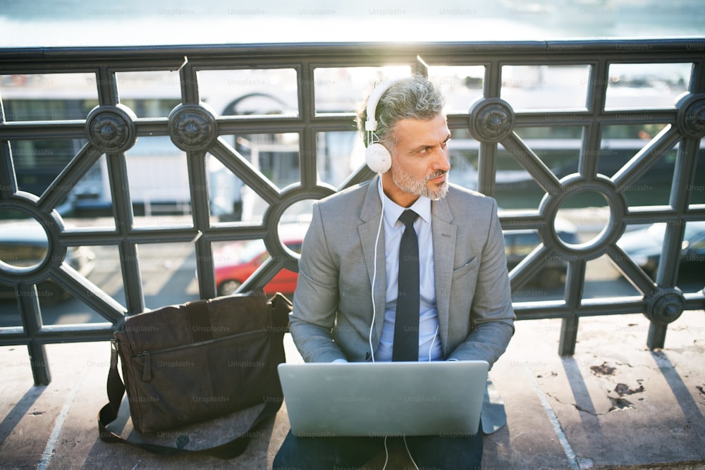 Handsome mature businessman with laptop in a city. Man sitting on a bridge, using laptop and headphones.