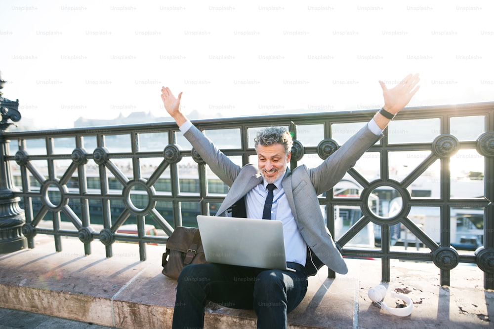 Handsome mature businessman with laptop in a city. Man sitting on a bridge, stretching arms.