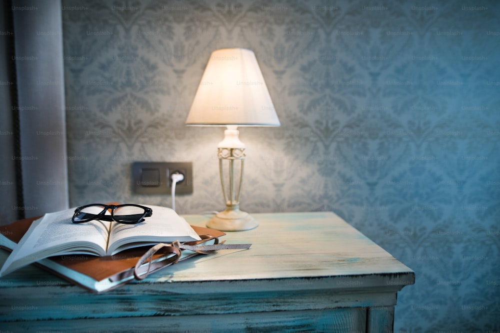 A lamp, a book and glasses on a bedside table in a hotel room.