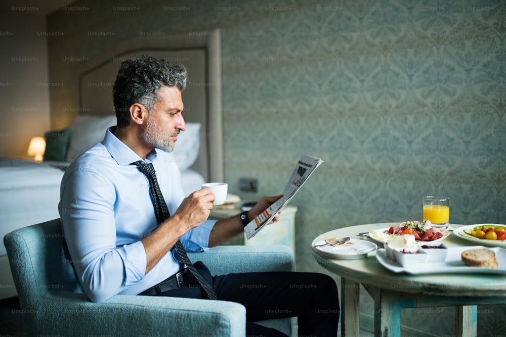 Empresário maduro e bonito tomando café da manhã em um quarto de hotel, lendo jornais e tomando café.