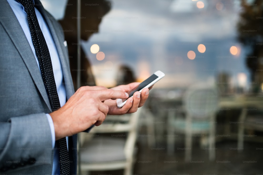 Hombre de negocios irreconocible con teléfono inteligente parado en la cafetería de un hotel al aire libre. Mensajes de texto del hombre.