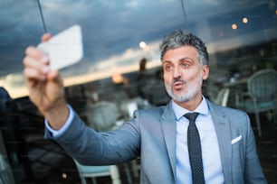 Hombre de negocios maduro con teléfono inteligente tomando una selfie en la cafetería de un hotel al aire libre.