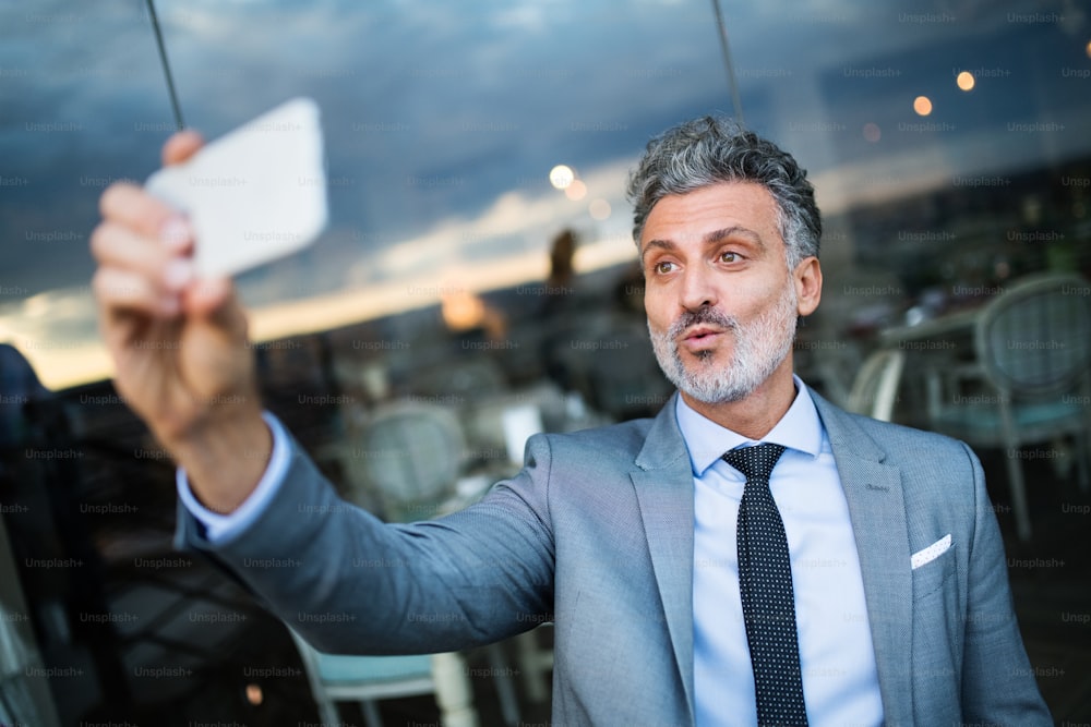 Mature businessman with smartphone taking a selfie in an outdoor hotel cafe.