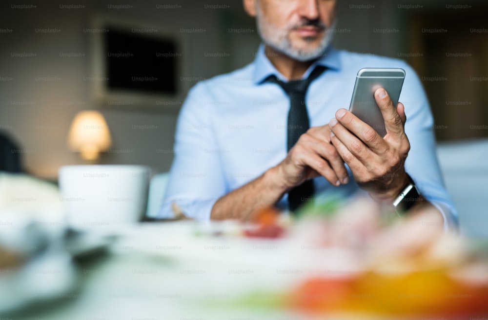 Unrecognizable mature businessman with smartphone in a hotel room. Man text messaging while having breakfast.