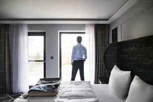 Bello uomo d'affari maturo in piedi alla finestra in una stanza d'albergo. Veduta posteriore.