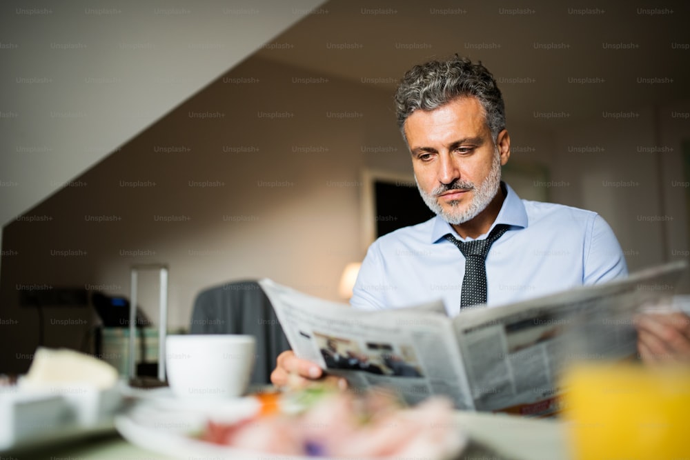Hombre de negocios maduro y guapo desayunando en una habitación de hotel, leyendo periódicos.