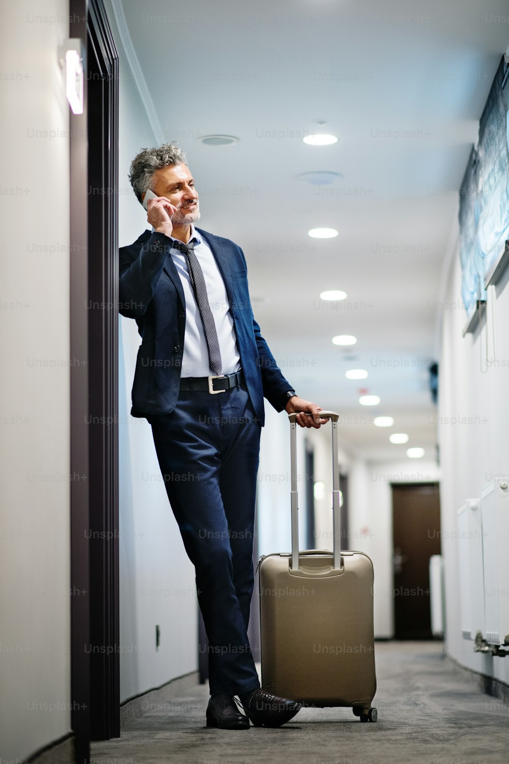 Hombre de negocios maduro con teléfono inteligente en el pasillo de un hotel. Hombre guapo parado en el pasillo de un hotel, haciendo una llamada telefónica.