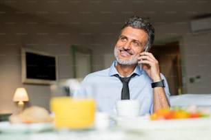 Mature, handsome businessman with smartphone in a hotel room. Man making a phone call while having breakfast.