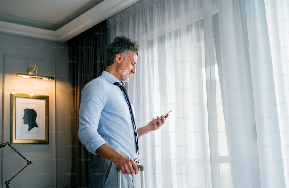 Mature businessman with smartphone in a hotel room. Handsome man standing at the window, texting.