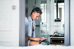 Mature businessman in a hotel room bathroom. Handsome man sitting on the toilet, using tablet.