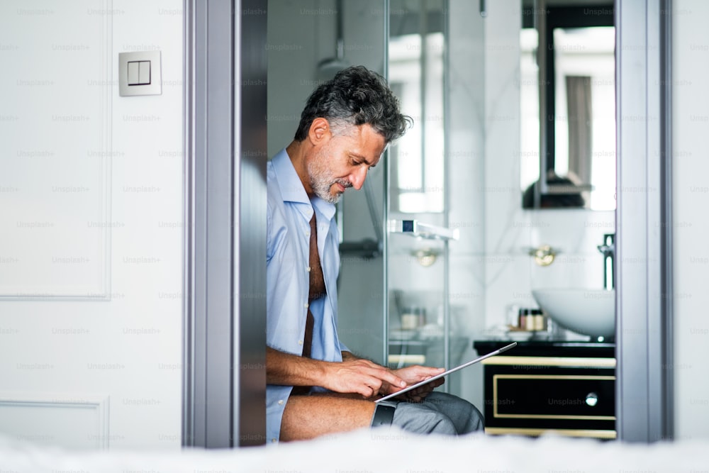 Homme d’affaires mature dans la salle de bain d’une chambre d’hôtel. Bel homme assis sur les toilettes, utilisant une tablette.