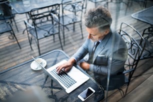 Reifer Geschäftsmann mit Laptop und Smartphone, der vor einem Café sitzt. Mann arbeitet an einem Laptop. Durch Glas geschossen.