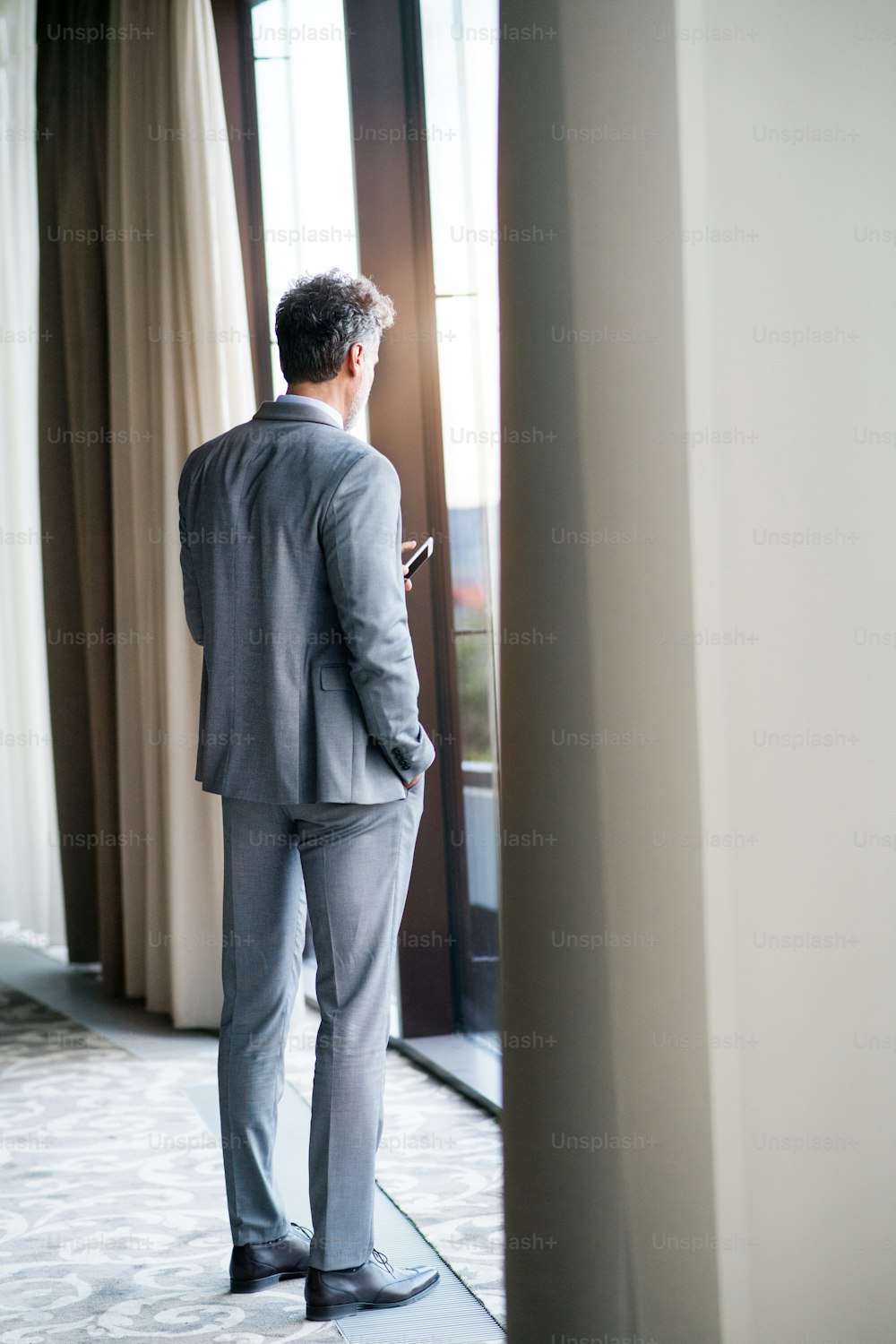 Hombre de negocios maduro en un hotel con un teléfono inteligente. Hombre haciendo de pie en la ventana, enviando mensajes de texto. Vista trasera.