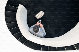 Businessman with smartphone and suitcase standing at the bottom of the staircase, texting. Top view.
