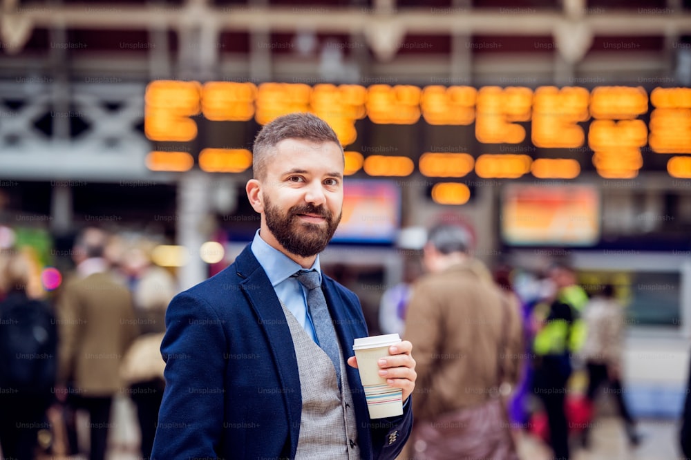 Hombre de negocios hipster sosteniendo una taza de café desechable en la concurrida estación de tren