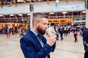 Hipster-Geschäftsmann mit einem Einwegbecher und Kaffee trinkend am überfüllten Bahnhof