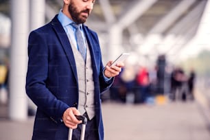 Uomo d'affari hipster in giacca e cravatta con lo smartphone, in attesa all'aeroporto, giornata di sole