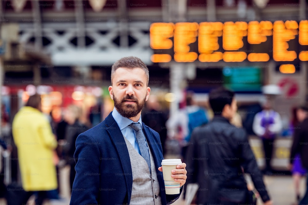 Hombre de negocios hipster sosteniendo una taza de café desechable en la concurrida estación de tren