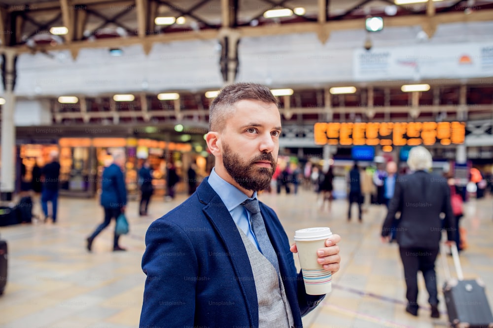 Uomo d'affari hipster che tiene una tazza di caffè usa e getta nella stazione ferroviaria affollata
