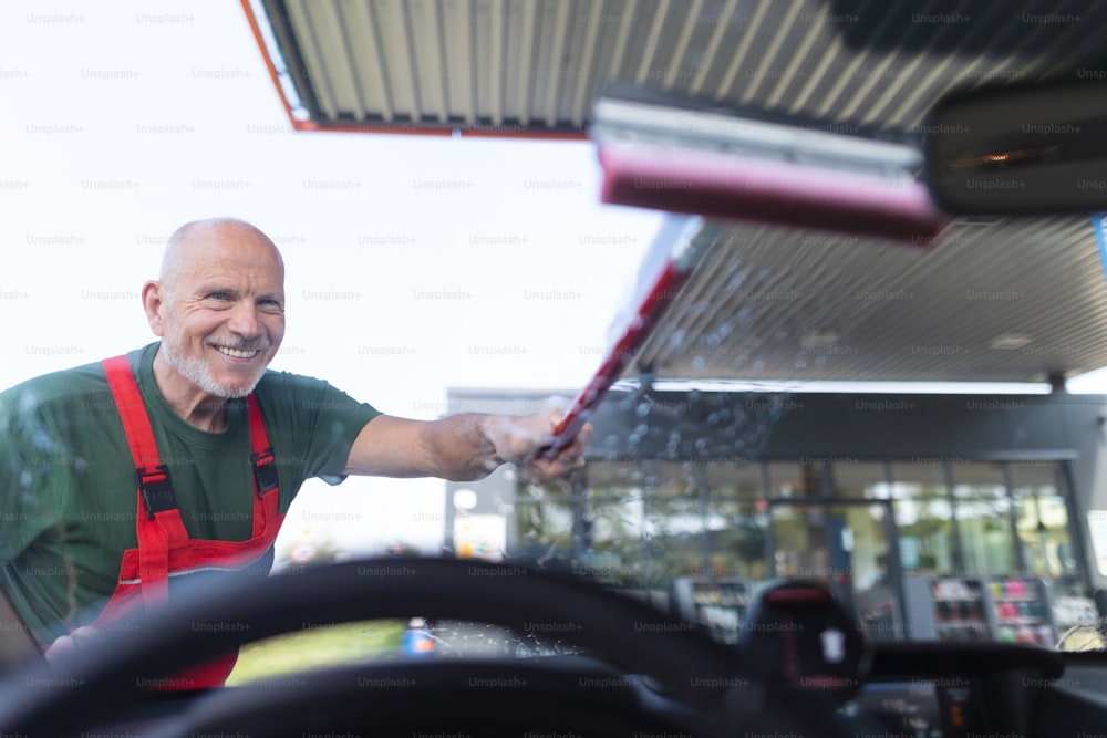 Cheerful senior worker washing a car window at gas station.