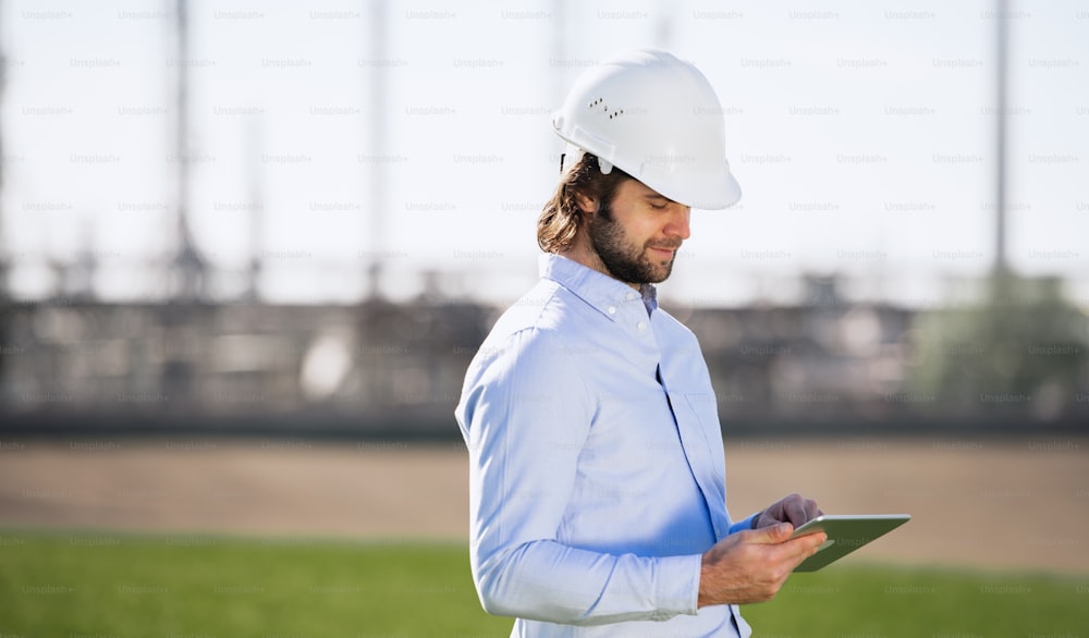 Jovem engenheiro com capacete em pé ao ar livre pela refinaria de petróleo, usando tablet. Espaço de cópia.