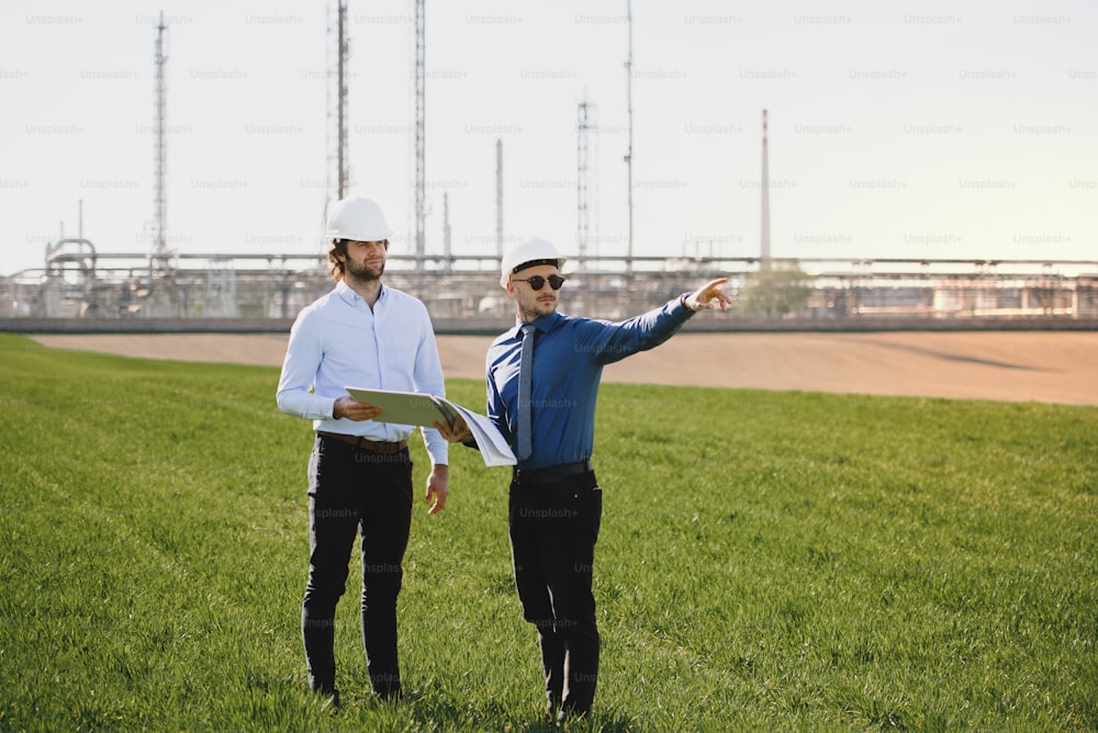 Deux jeunes ingénieurs debout à l’extérieur près d’une raffinerie de pétrole, discutant de problèmes. Espace de copie.