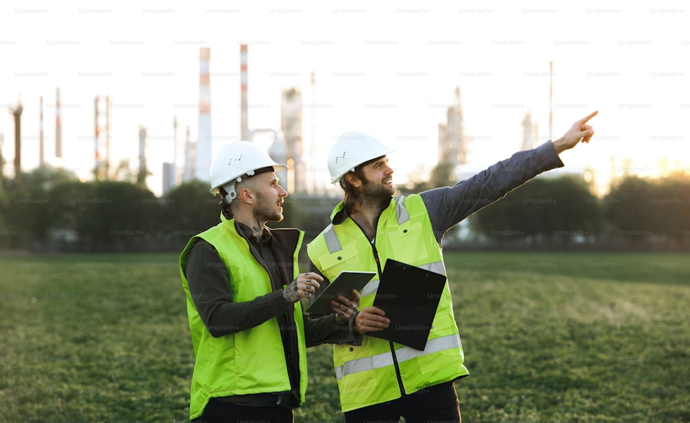 Vista frontal de dos jóvenes ingenieros con tabletas de pie al aire libre junto a una refinería de petróleo, discutiendo temas.