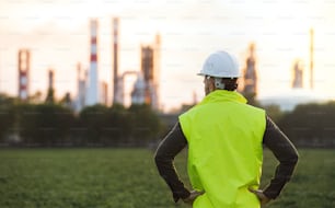 Rear view of young engineer standing outdoors by oil refinery, arms on hips. Copy space.