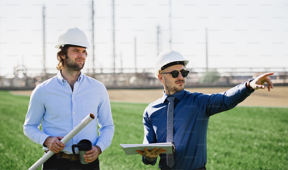 Two young engineers with hard hat standing outdoors by oil refinery, discussing issues. Copy space.