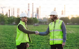 Two young engineers with hard hat standing outdoors by oil refinery, shaking hands.