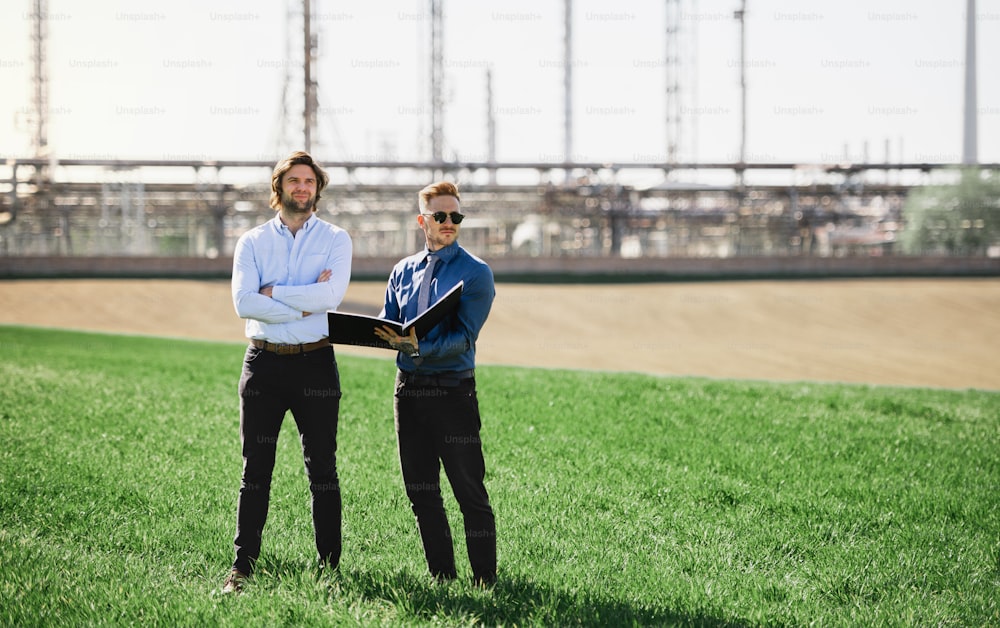 Two young engineers standing outdoors by oil refinery, discussing issues. Copy space.