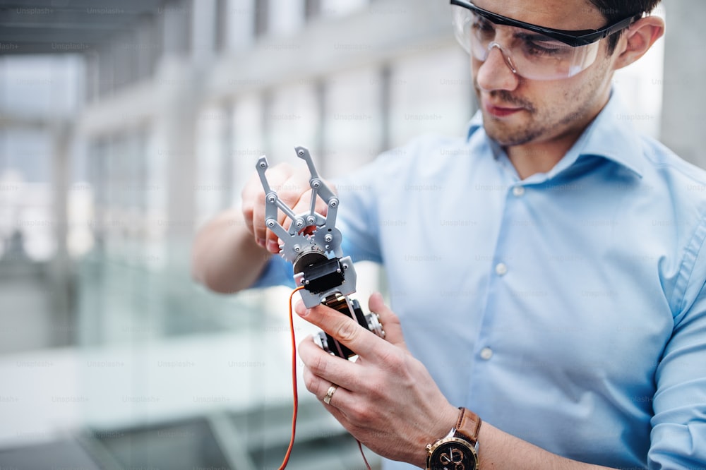A young businessman or scientist with robotic hand and safety glasses standing in office, working.