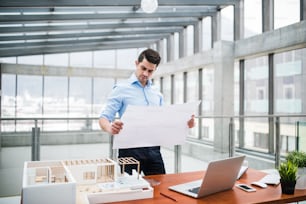 Young businessman or architect with model of a house and blueprints standing at the desk in office, working.