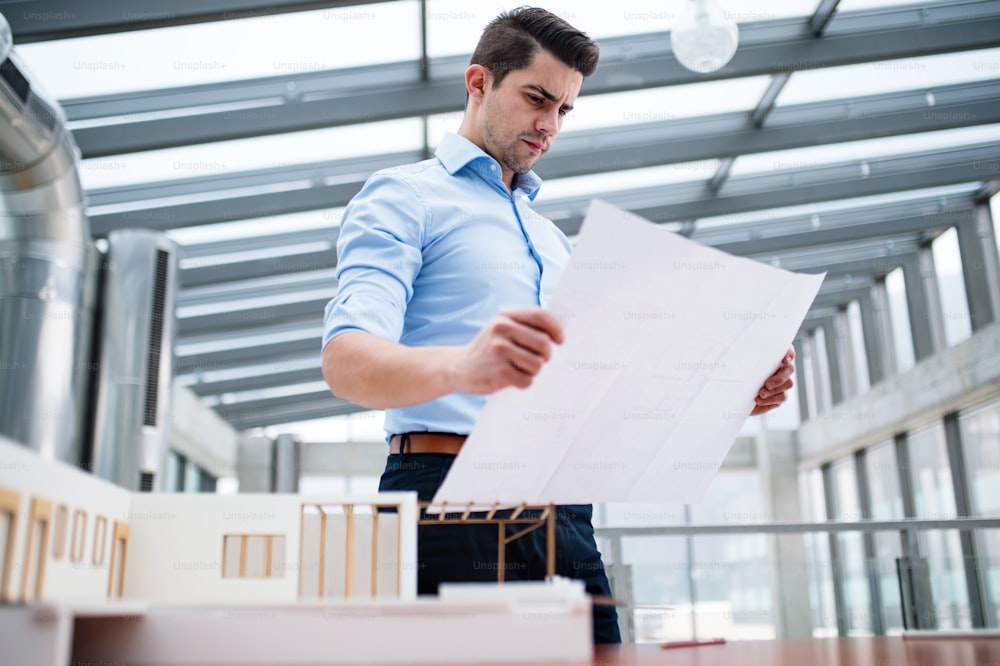 Young businessman or architect with model of a house standing at the desk in office, looking at blueprints when working.