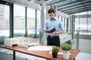 Giovane uomo d'affari o architetto con modello di una casa in piedi alla scrivania in ufficio, prendendo appunti quando si lavora.