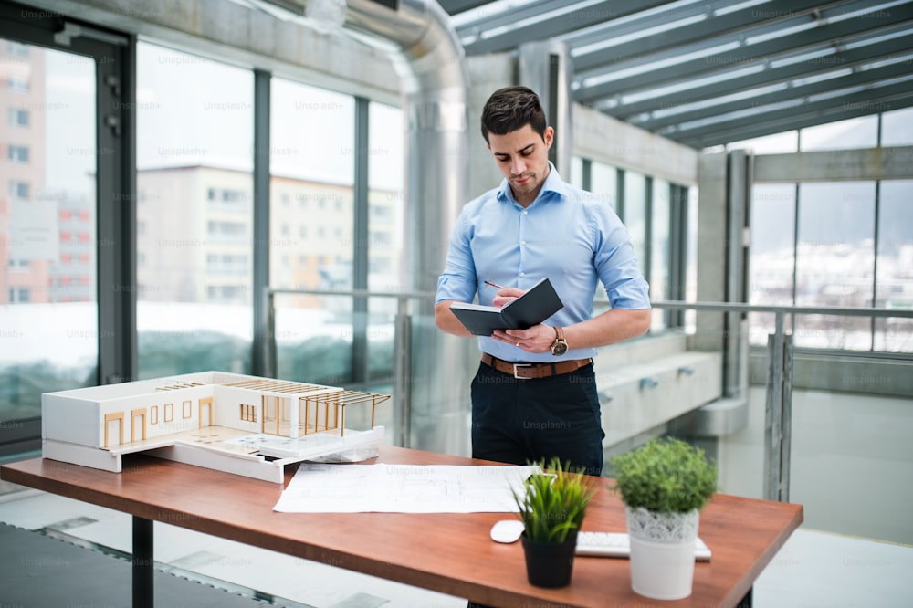 Young businessman or architect with model of a house standing at the desk in office, making notes when working.