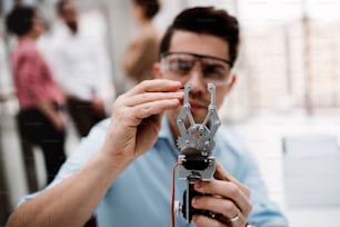 Un jeune homme d’affaires ou scientifique avec des mains robotisées et des lunettes de sécurité debout dans le bureau, en train de travailler.
