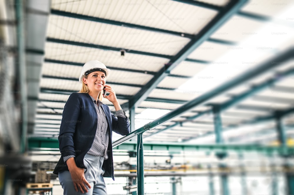 Ein Porträt einer glücklichen Industrieingenieurin mit Smartphone, die in einer Fabrik steht und telefoniert.