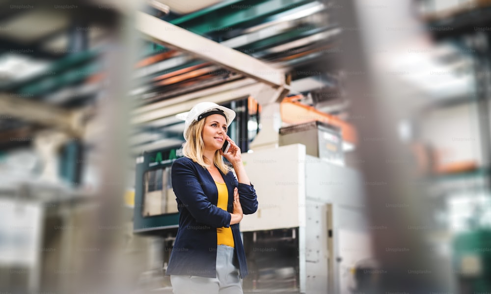 Ein Porträt einer glücklichen Industrieingenieurin am Telefon, die in einer Fabrik steht.