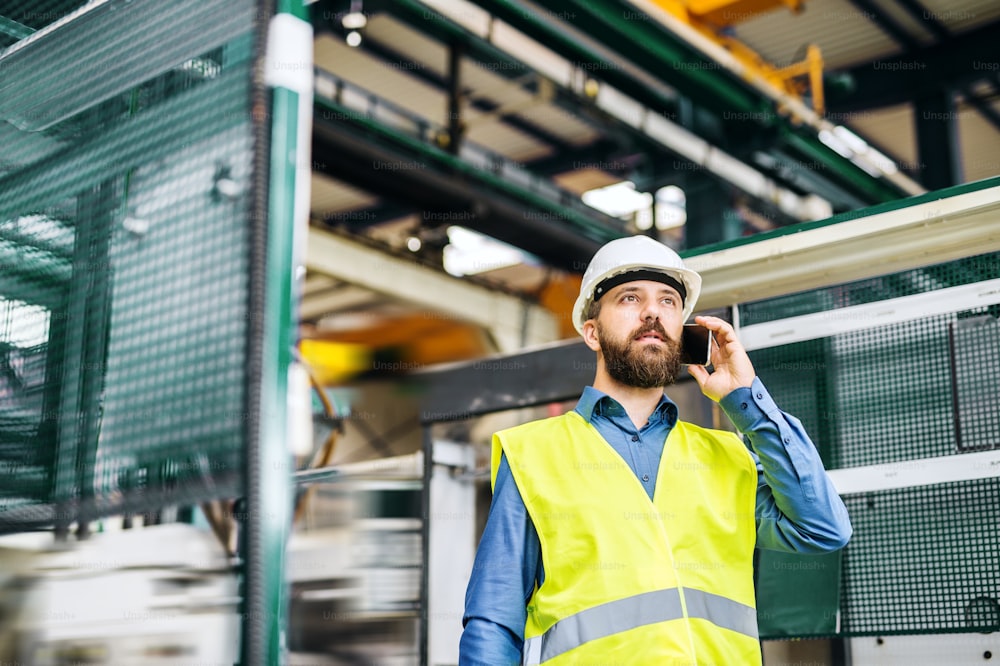 Un ritratto di un maturo ingegnere industriale con smartphone che lavora in una fabbrica, facendo una telefonata.