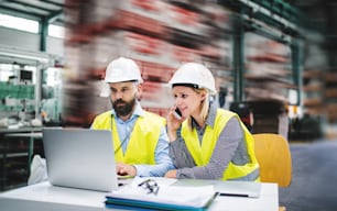 A portrait of a mature industrial man and woman engineer with laptop and smartphone in a factory, working.
