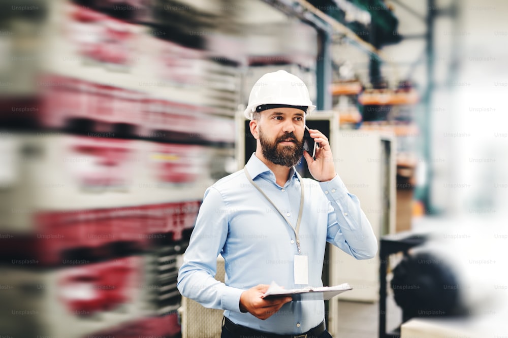 A portrait of a mature industrial man engineer with clipboard and smartphone in a factory, making a phone call. Copy space.