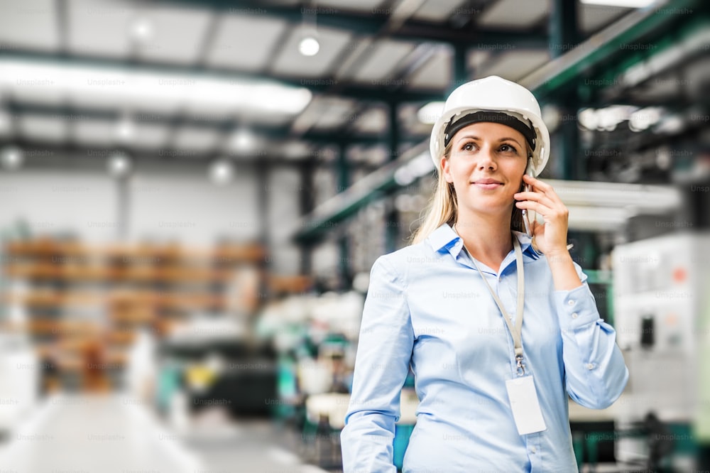 Portrait d’une jeune ingénieure industrielle avec smartphone et casque blanc debout dans une usine, faisant un appel téléphonique. Espace de copie.