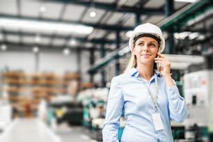 Portrait d’une jeune ingénieure industrielle avec smartphone et casque blanc debout dans une usine, faisant un appel téléphonique. Espace de copie.